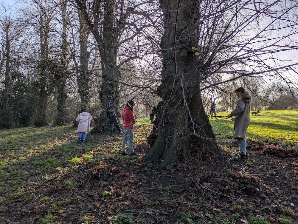 Three young people standing by some large trees. It is winter, the branches are bare and the people wear coats