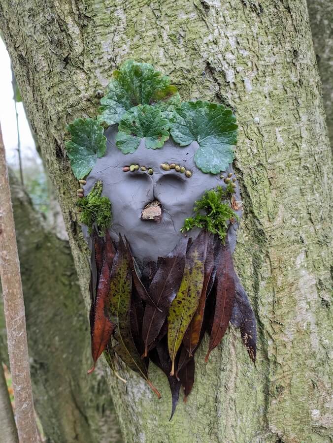 A clay face sculpted onto a tree. Leaves are stuck on to make facial features