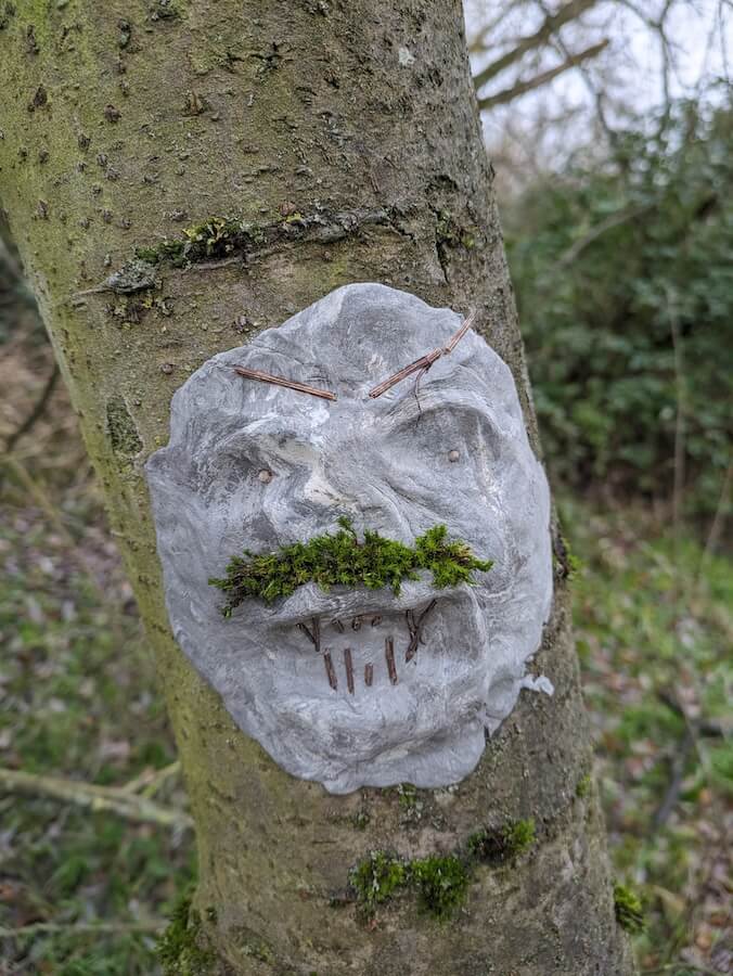 A clay face sculpted onto a tree. It has a moss moustache and twigs for teeth